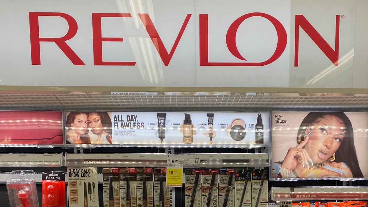 A large "Revlon" sign in red letters above a wall of cosmetics products in a drugstore.