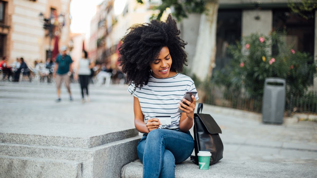 An online shopper is sitting outside on the street and is ordering something on her mobile phone with a credit card in hand.