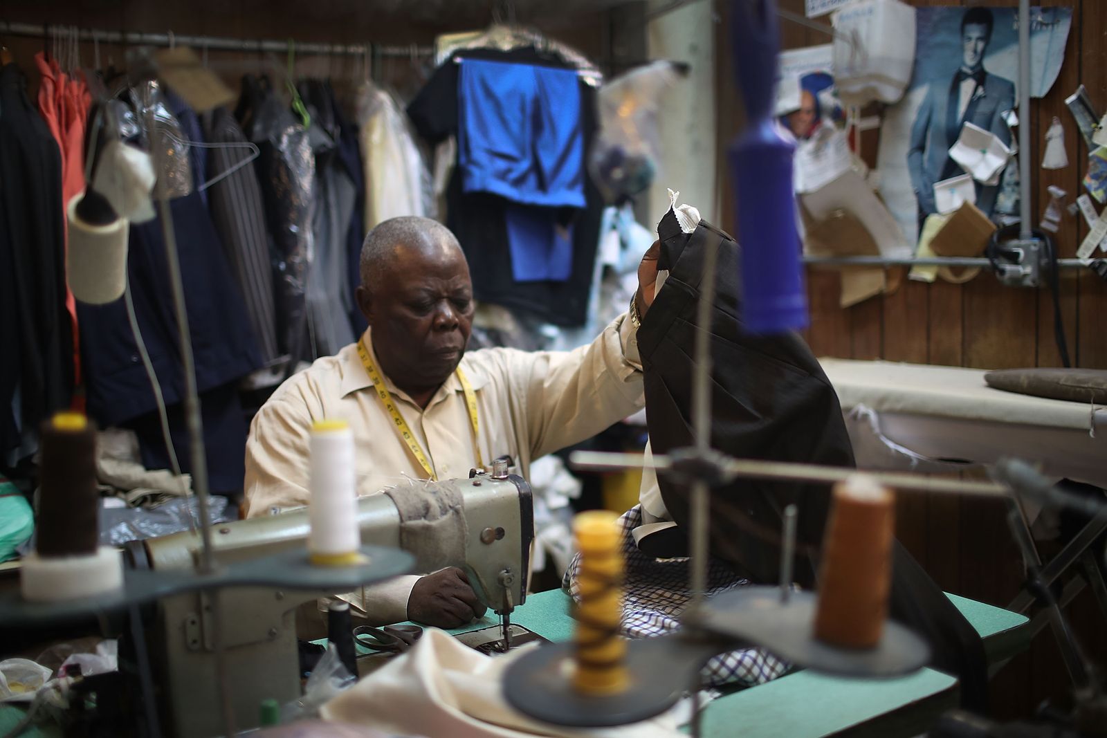 A man is sewing garments in a room surrounded by thread and apparel.